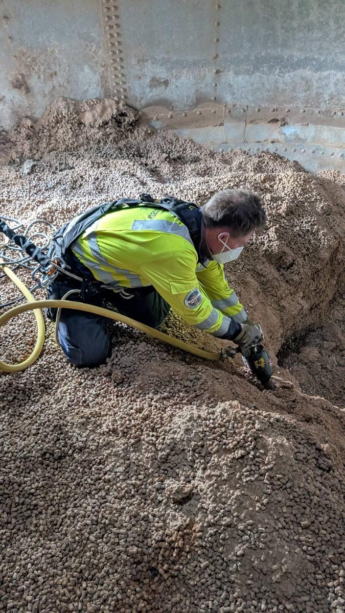 A QRF operative in harness and dust mask, kneeling on feed pellets inside a silo, breaking up compacted product with an air pick.