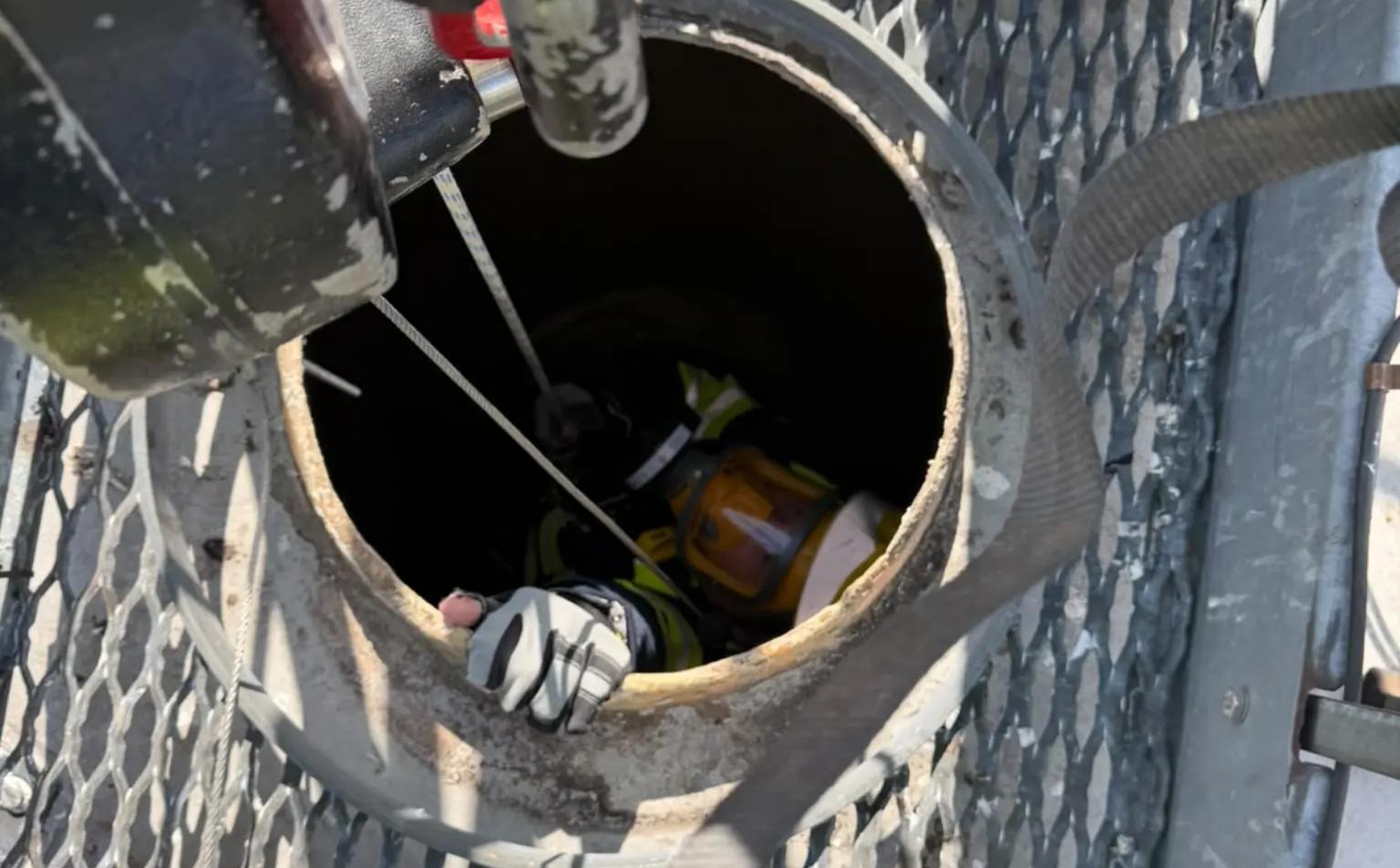 Looking down through a silo hatch at a QRF entrant in a respirator, on a winch line, with the rescue tripod and attendant above.