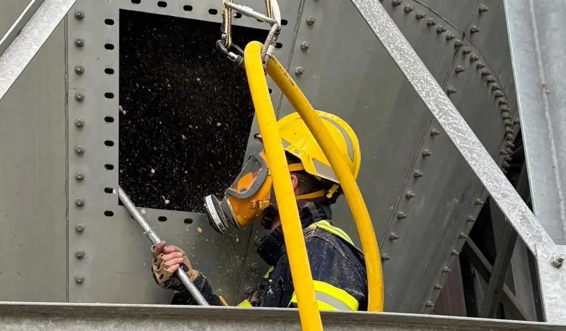 A QRF operative in a full face respirator clearing product through a silo hatch with an air lance, clipped to an anchor above.